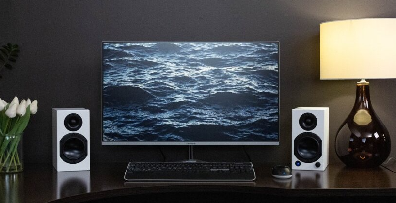 Lifestyle shot showcasing the Dayton Audio M4W speaker pair arranged on either side of a PC monitor, on top of a home office desk.